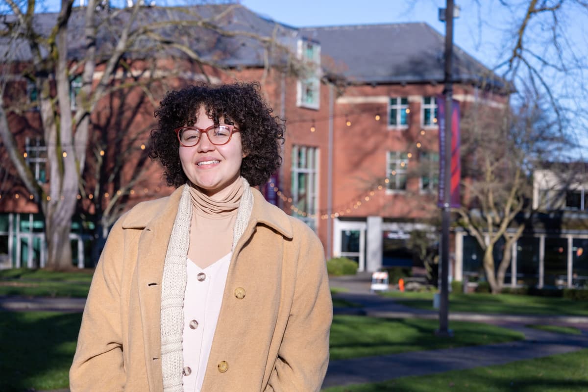 Photo of Esther in tan coat on campus with Eaton Hall in the background.