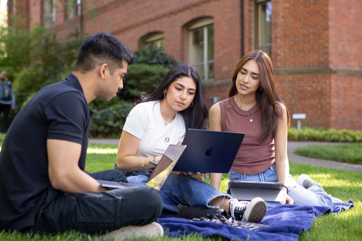 One male student and two female students sitting next to each other on the lawn looking at something on a laptop computer.
