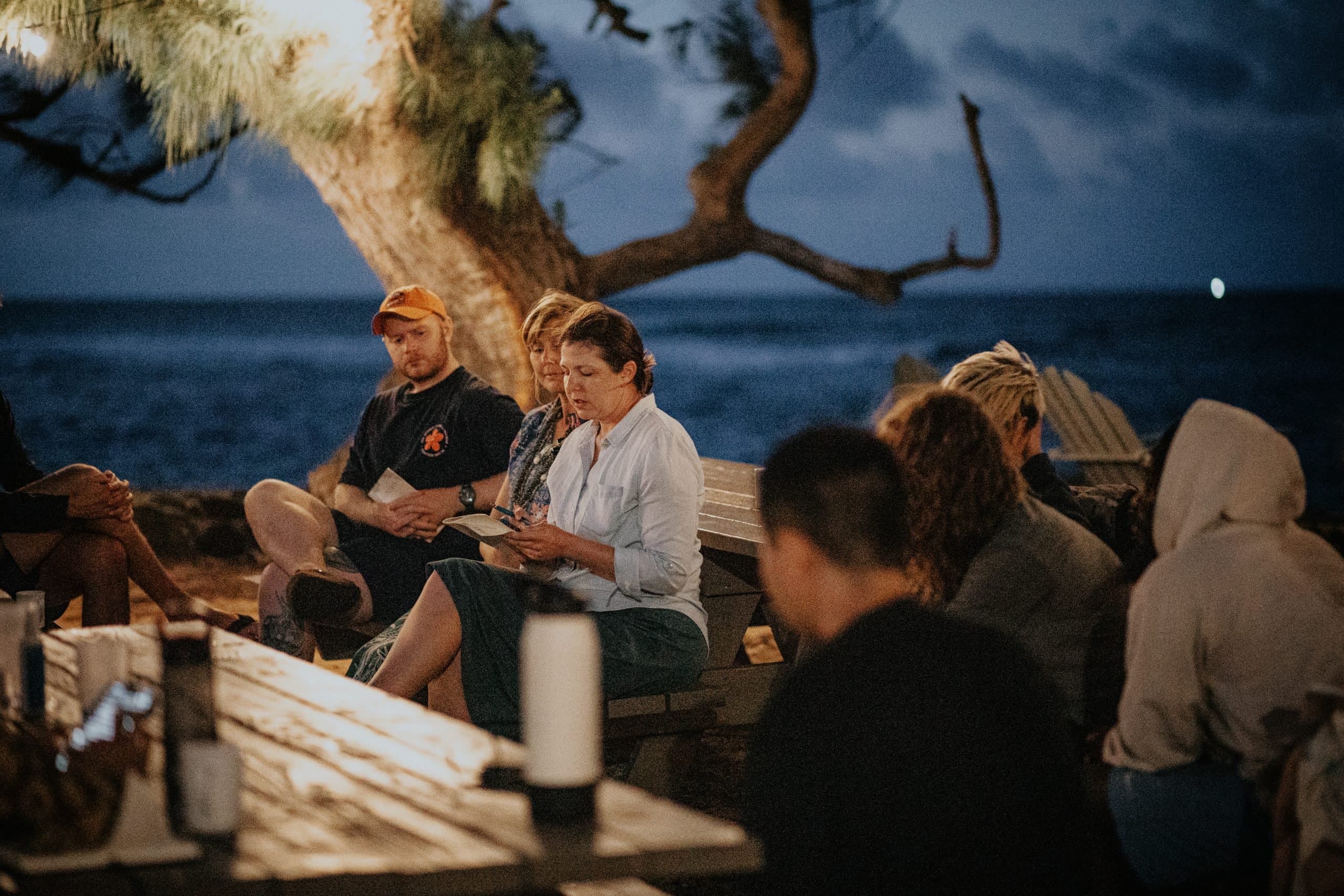 A person is reading to a small group of people sitting outside under a big tree at dusk.