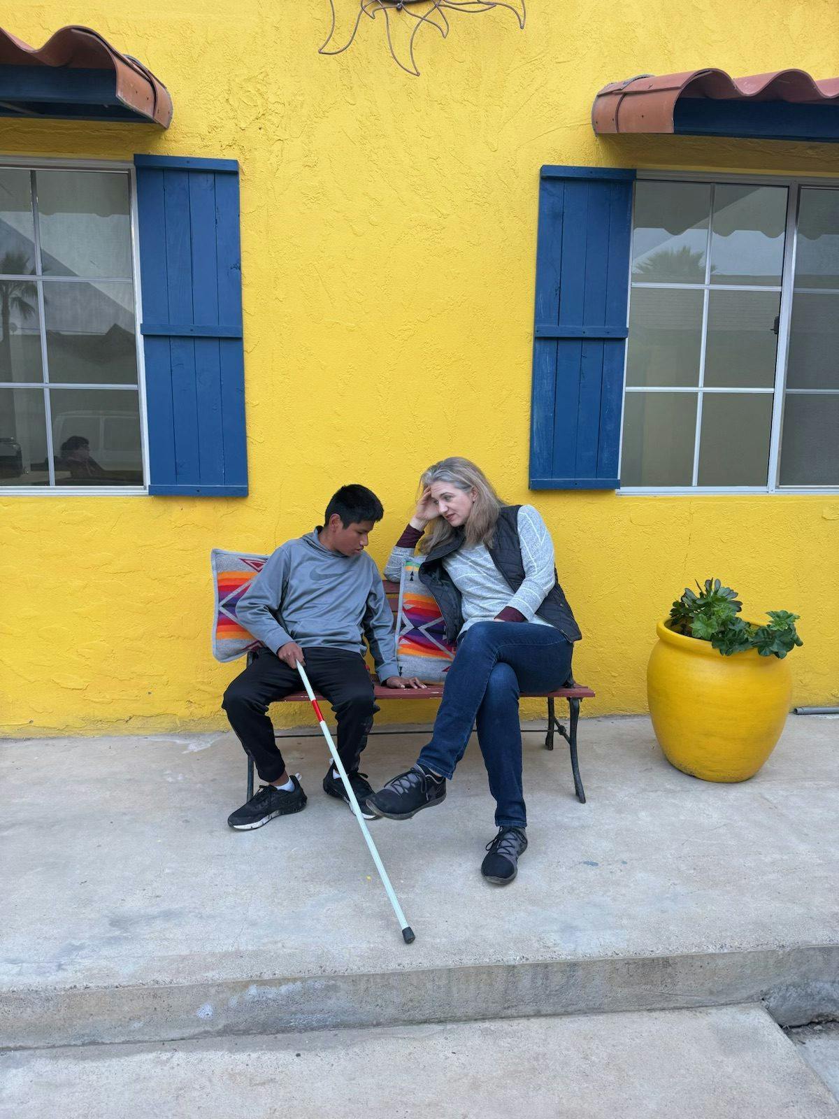 young man with disabilities sits on a bench next to a woman, Nicole Zinn, against a bright yellow building with windows with blue shutters in Mexico