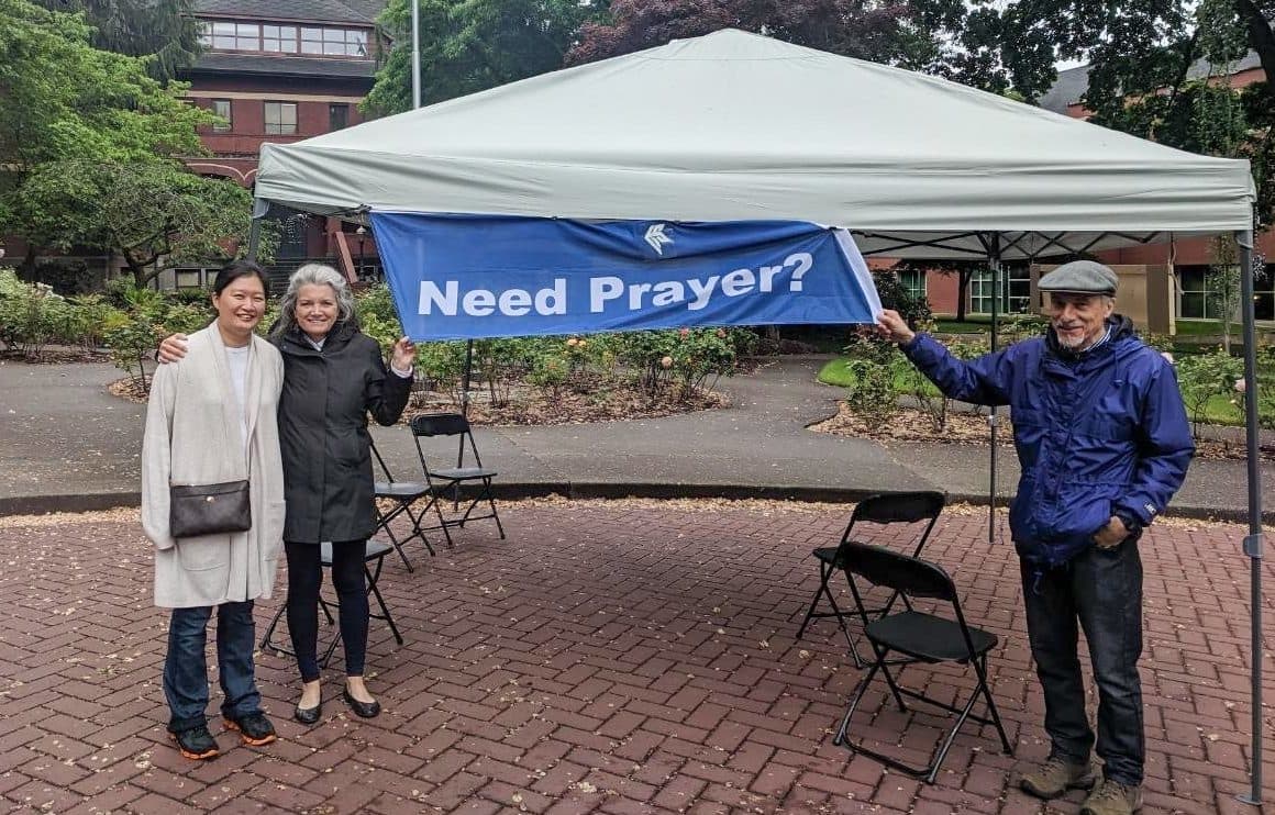 Three people standing under a sign that says "Need Prayer."